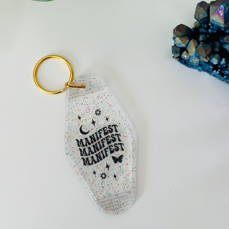 May include: A clear plastic motel key tag keychain with glitter and the words "MANIFEST" repeated three times in black. A crescent moon, stars, and a butterfly are also printed on the tag.