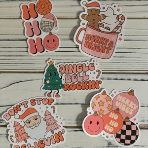 May include: Set of five Christmas-themed stickers featuring illustrations of Santa Claus, a gingerbread man, a Christmas tree, and ornaments. The stickers have a retro, vintage aesthetic with pink, orange, and white colors. The text on the stickers includes "Ho Ho Ho", "Merry & Bright", "Jingle Bell Rockin", "Don't Stop Believin", and "Merry Vibes".