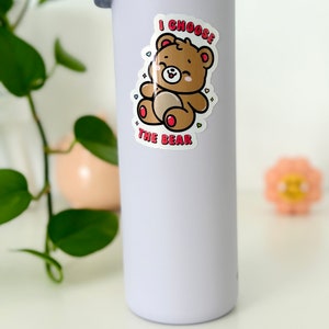 Może przedstawiać: Biała butelka z wodą z naklejką przedstawiającą kresk&oacute;wkowego misia. Naklejka głosi "I choose the bear" z sercami wok&oacute;ł misia.
