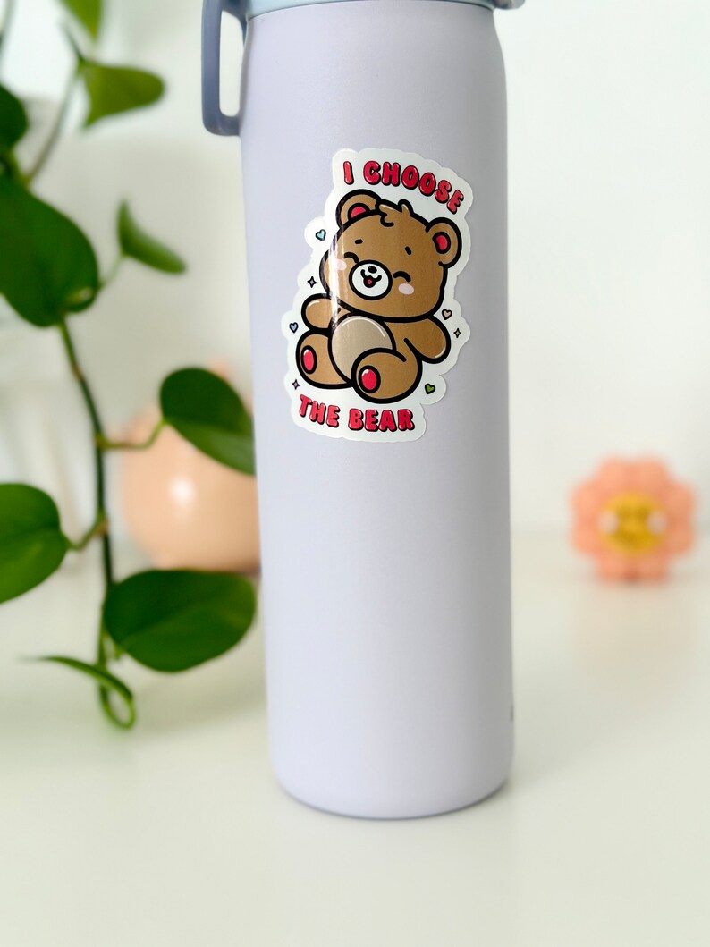 Może przedstawiać: Biała butelka z wodą z naklejką przedstawiającą kresk&oacute;wkowego misia. Naklejka głosi "I choose the bear" z sercami wok&oacute;ł misia.