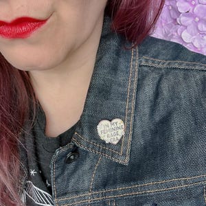 May include: A person wearing a denim jacket with a heart-shaped pin that says "In My Feminine Rage Era".