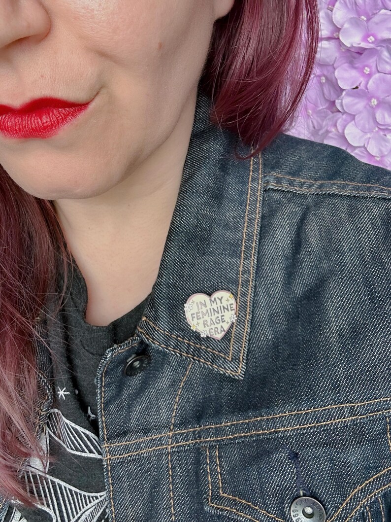 May include: A person wearing a denim jacket with a heart-shaped pin that says "In My Feminine Rage Era".