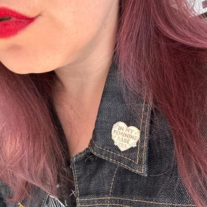 May include: A gold heart-shaped pin with the text "In My Feminine Rage" in white lettering. The pin is worn on a denim jacket.