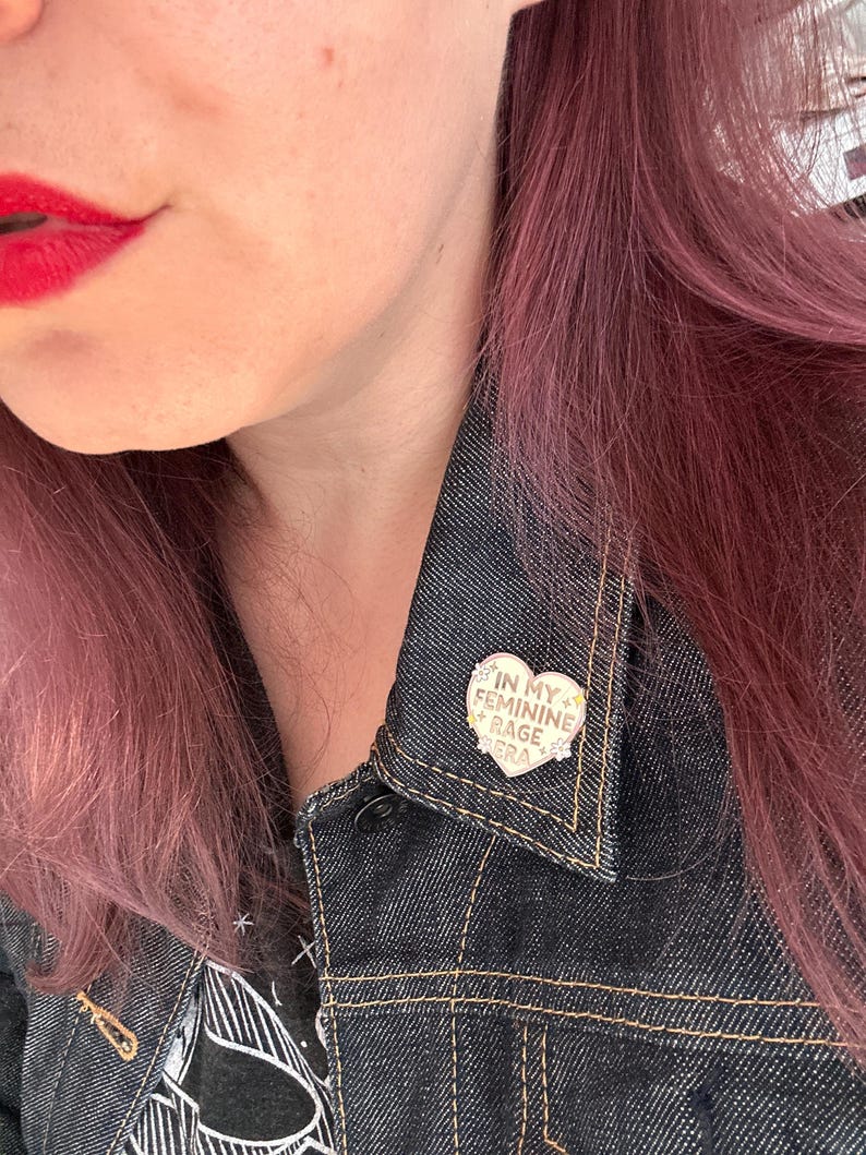 May include: A gold heart-shaped pin with the text "In My Feminine Rage" in white lettering. The pin is worn on a denim jacket.
