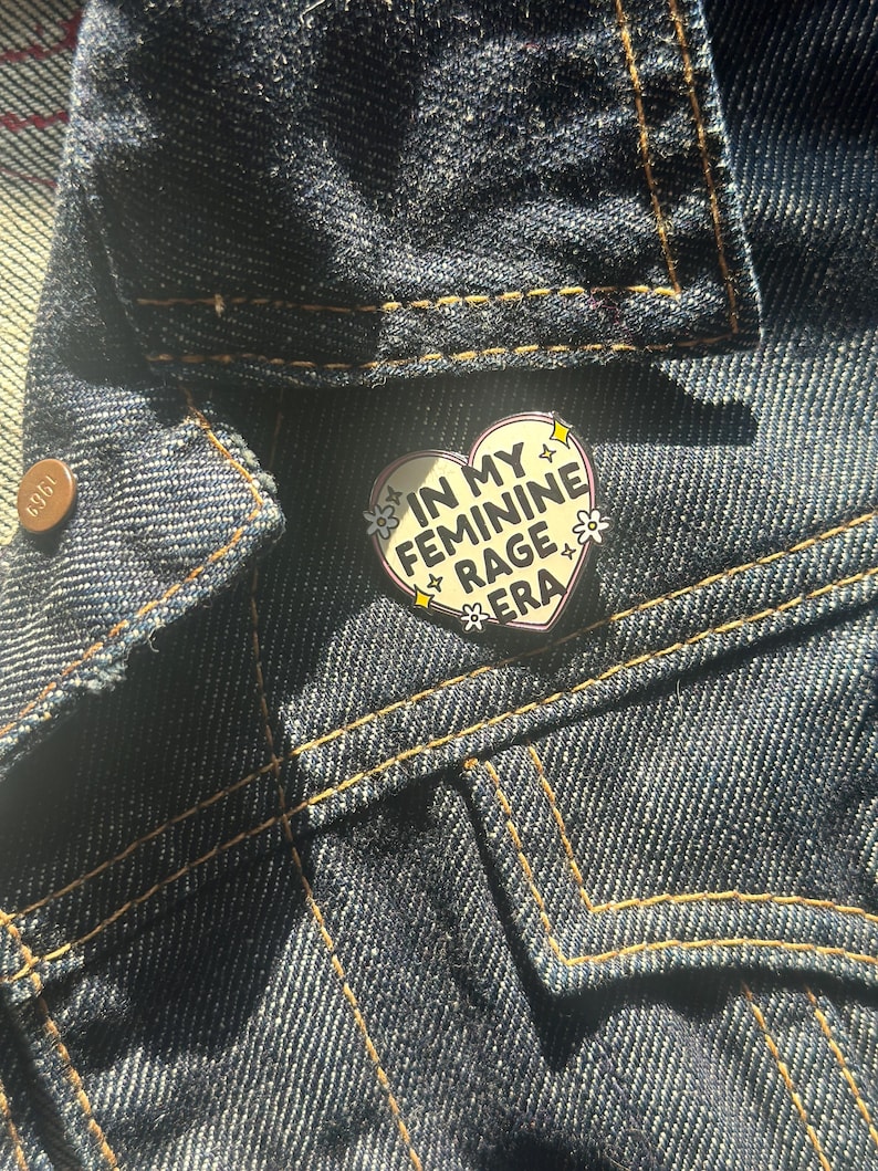 May include: A white heart-shaped enamel pin with a black outline. The pin says 'In My Feminine Rage Era' in black text with white flowers around the text.