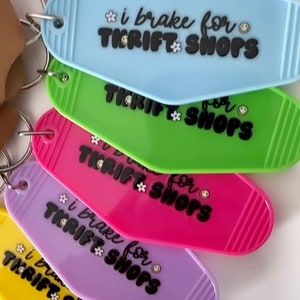 May include: A set of six colorful keychains shaped like motel room keys. Each keychain has the text "i brake for thrift shops" with a flower and smiley face graphic. The keychains are in the colors blue, green, pink, purple, yellow, and orange.