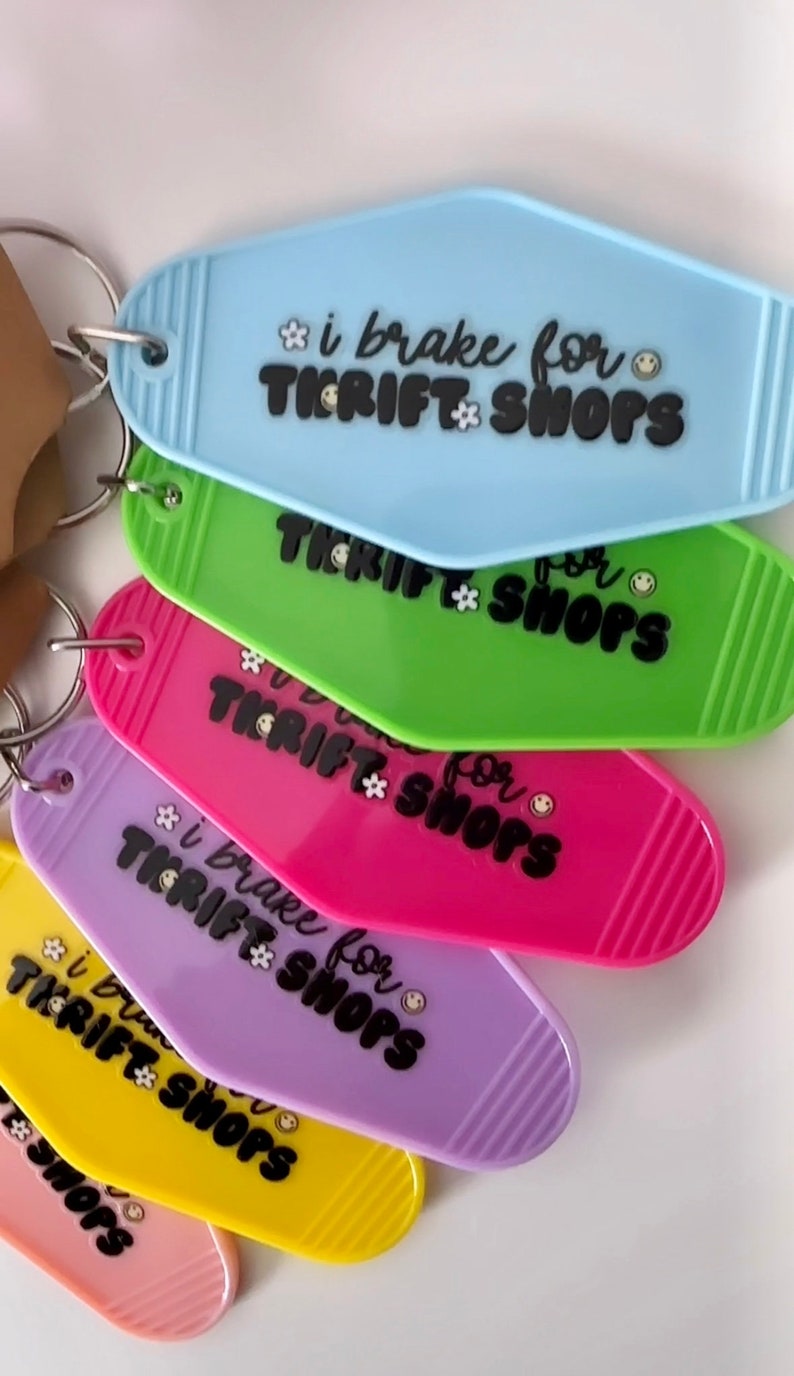 May include: A set of six colorful keychains shaped like motel room keys. Each keychain has the text "i brake for thrift shops" with a flower and smiley face graphic. The keychains are in the colors blue, green, pink, purple, yellow, and orange.