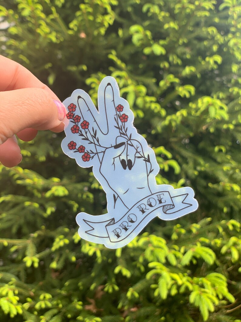 K&ouml;nnte beinhalten: Ein blauer Aufkleber mit einer Hand, die ein Peace-Zeichen macht und einen Zweig mit roten Blumen h&auml;lt. Der Aufkleber hat ein Banner mit der Aufschrift "PRO ROE" in schwarzen Buchstaben.