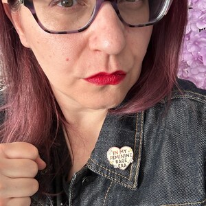 May include: A person wearing a denim jacket with a heart-shaped pin that says "In My Feminine Rage Era".