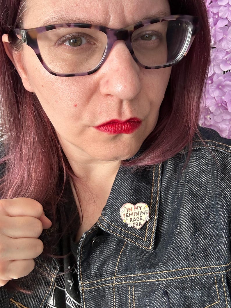 May include: A person wearing a denim jacket with a heart-shaped pin that says "In My Feminine Rage Era".