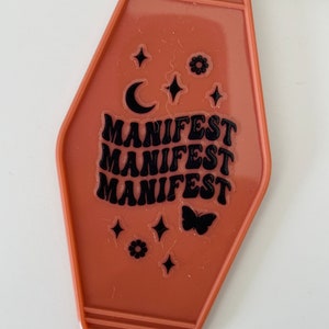 May include: A brown motel key tag keychain with the word "MANIFEST" repeated three times in black lettering. The keychain also features a crescent moon, stars, and a butterfly.