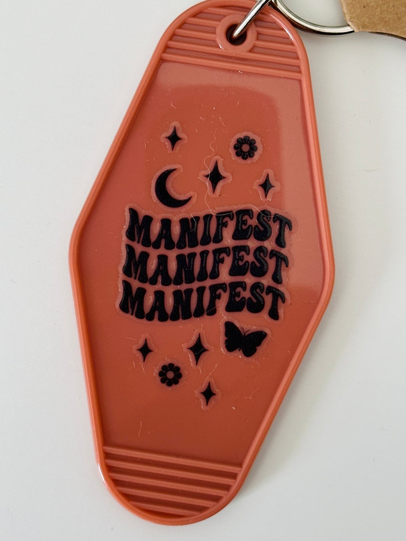 May include: A brown motel key tag keychain with the word "MANIFEST" repeated three times in black lettering. The keychain also features a crescent moon, stars, and a butterfly.