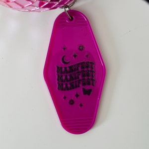 May include: A pink plastic motel key tag with the word "MANIFEST" repeated four times in black text. The key tag also features a crescent moon, stars, and a butterfly.