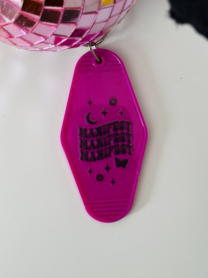 May include: A pink plastic motel key tag with the word "MANIFEST" repeated four times in black text. The key tag also features a crescent moon, stars, and a butterfly.