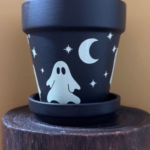 May include: Black terracotta planter with a white ghost, crescent moon, and stars design. The planter has a matching saucer.