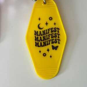 May include: Yellow motel key tag with black text that reads "MANIFEST" three times. The tag is decorated with a crescent moon, stars, a flower, and a butterfly.