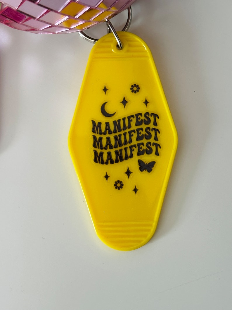 May include: Yellow motel key tag with black text that reads "MANIFEST" three times. The tag is decorated with a crescent moon, stars, a flower, and a butterfly.