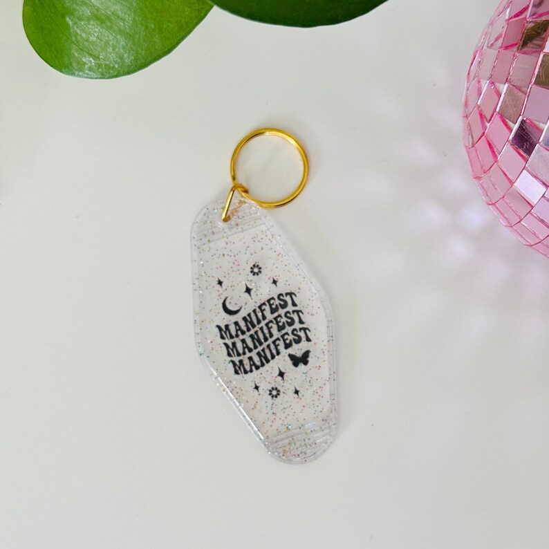 May include: A clear, glittery keychain with a gold ring. The keychain is shaped like a motel key and has the word "MANIFEST" repeated three times in black text. There is a small crescent moon and a butterfly graphic on the keychain.