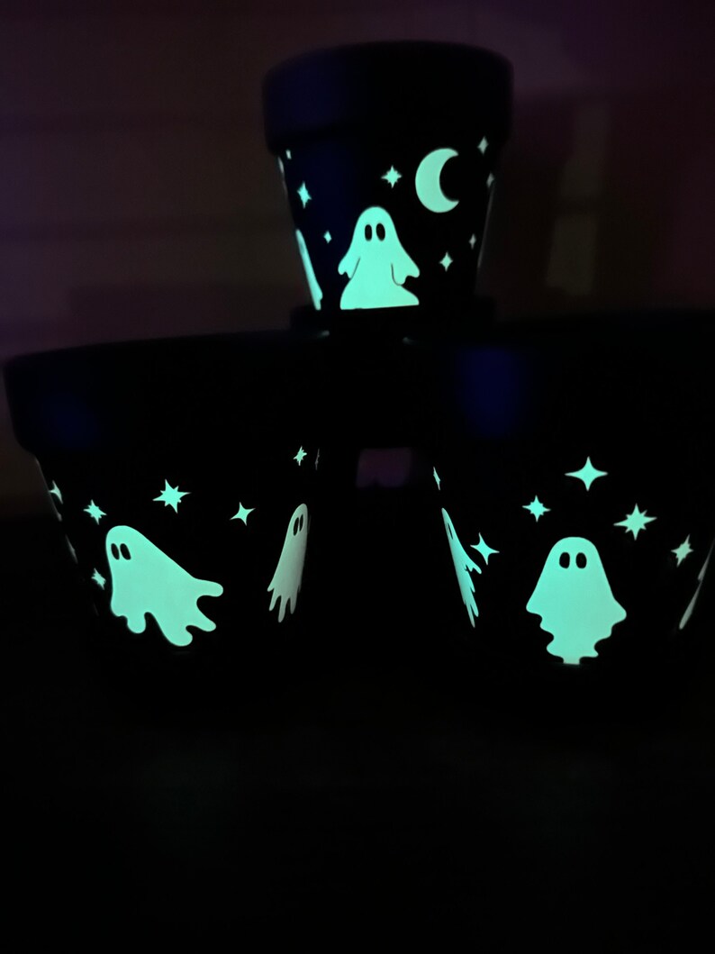 May include: Three black terracotta flower pots with glow-in-the-dark paint. The pots are decorated with white stars, a crescent moon, and ghosts.