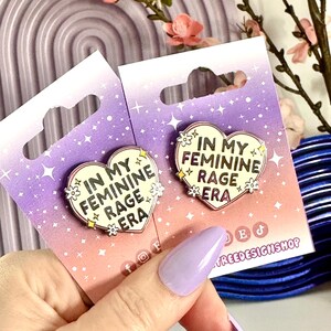 May include: Two enamel pins with a heart shape and the text "In My Feminine Rage Era" on a purple background with white stars. The pins are on a card with the text "Treedesignshop" on the bottom right corner.