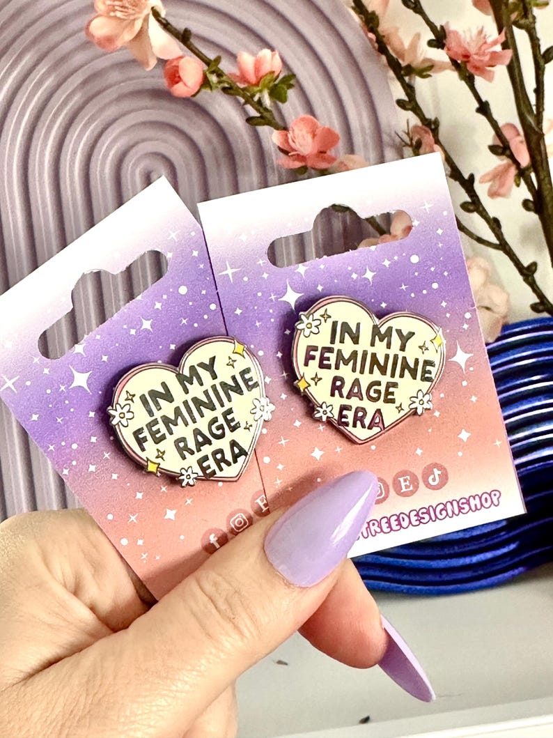 May include: Two enamel pins with a heart shape and the text "In My Feminine Rage Era" on a purple background with white stars. The pins are on a card with the text "Treedesignshop" on the bottom right corner.