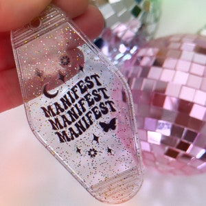 May include: A clear plastic keychain with glitter embedded in the plastic. The keychain is shaped like a motel key and has the word "MANIFEST" repeated three times in black lettering. There is a small butterfly and stars on the keychain.