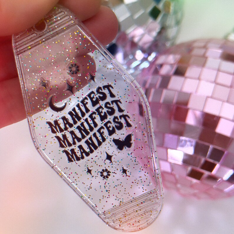 May include: A clear plastic keychain with glitter embedded in the plastic. The keychain is shaped like a motel key and has the word "MANIFEST" repeated three times in black lettering. There is a small butterfly and stars on the keychain.