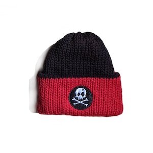 May include: A black and red knit beanie hat with a skull and crossbones patch. The hat is two-toned, with a black upper section and a red lower section. The patch is circular, with a white skull and crossbones on a black background.