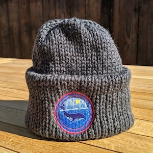 May include: A dark gray knit beanie with a folded brim. The beanie features a circular patch with a whale illustration, a blue ocean, a yellow sun, and a red border. The hat is made of a thick, textured yarn.