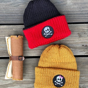 May include: Two knitted beanies, one black and red, the other mustard yellow, each with a skull and crossbones patch. A rolled-up felt item with a leather belt is also present. The beanies are likely for children or adults.
