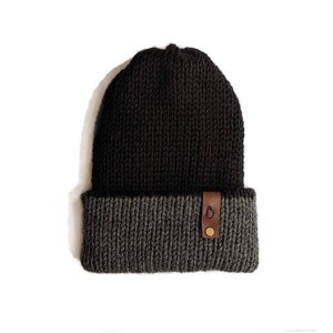 May include: A two-tone knit beanie in black and charcoal gray. The hat features a folded brim and a small brown leather tag with a logo. The beanie is made of a thick, warm material.