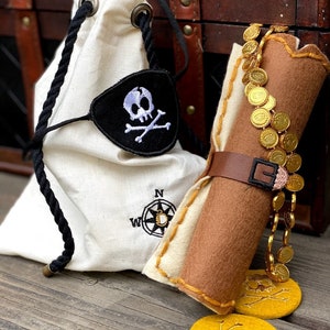 May include: A pirate-themed set featuring a white drawstring bag with a skull and crossbones eye patch, a rolled-up scroll with a leather strap, and gold coin decorations. The bag has a compass design. The set is on a wooden surface.