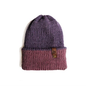 May include: A knitted beanie in shades of purple and mauve. The hat features a folded brim and a small brown leather tag with a logo. The beanie is made of a thick, textured yarn.