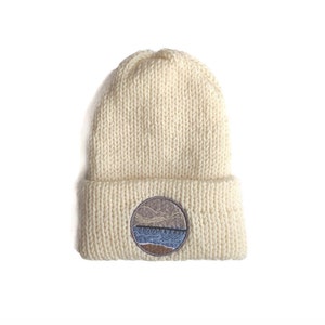 May include: Cream-colored knit beanie with a folded brim. A circular patch on the front features a landscape design with mountains, a body of water, and a shoreline. The hat is made of a thick, textured yarn.