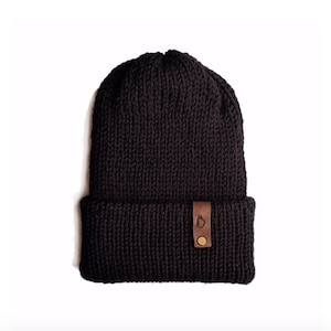 May include: A dark brown knit beanie with a folded brim. A small, rectangular brown leather tag is sewn onto the brim, with a gold-colored button and a small logo. The hat is made of a thick, textured yarn.