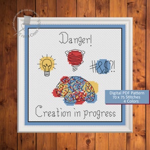 Puede incluir: Un patrón de punto de cruz digital que presenta un cerebro con un esquema de color rojo, amarillo y azul. El texto "Danger!" está por encima del cerebro, y el texto "Creation in progress" está por debajo del cerebro. El patrón es de 70 x 75 puntos y utiliza 4 colores.