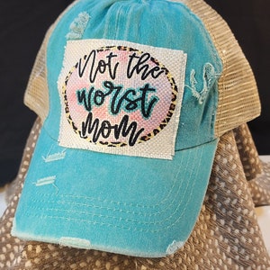 May include: A distressed turquoise baseball cap with a tan mesh back. The front patch reads "Not the worst Mom" in black script over a watercolor and leopard print design. The hat has a pre-worn look.