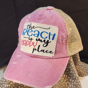 May include: Pink distressed baseball cap with a tan mesh back. The front of the cap features a patch with the text "the beach is my happy place" in black, blue, and red lettering.