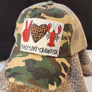 Può includere: Cappellino da baseball mimetico con retro in rete. La toppa anteriore presenta un segno di pace, un cuore con stampa leopardata e un gambero rosso, con le parole "PEACE LOVE CRAWFISH" sotto. Il cappellino ha un aspetto usurato.