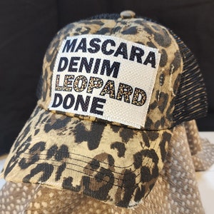 May include: A leopard print trucker hat with a black mesh back. The front patch features the text "Mascara Denim Leopard Done" in black and leopard print. The hat has a curved peak and a button on top.
