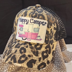 Puede incluir: Gorra de béisbol con estampado de leopardo y parte trasera de malla negra. El parche frontal presenta una caravana rosa y blanca con las palabras "Happy Camper" bordadas en negro.