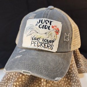 Op de afbeelding: Grijze truckerpet met een versleten look, mesh achterkant en gebogen klep. De pet heeft een patch met een illustratie van een haan en de tekst "JUST A GIRL WHO LOVES PECKERS".