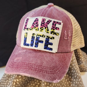 May include: A distressed maroon baseball cap with a mesh back. The front features a white patch with the words "LAKE LIFE" in a mix of leopard print and blue lettering. The cap has a pre-curved bill.