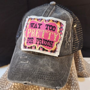 Peut inclure: Casquette de camionneur grise délavée avec un dos en filet noir. L'avant présente un écusson avec le texte "WAY TOO PRETTY FOR PRISON" en lettres roses et dorées. La casquette a un aspect usé avec des détails vieillis.