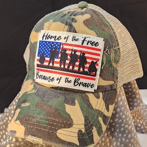 Pode incluir: Boné de beisebol camuflado com costas em malha. O patch frontal apresenta um design da bandeira americana com silhuetas de soldados e o texto "Home of the Free Because of the Brave."
