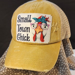 Puede incluir: Gorra de camionero de color amarillo mostaza con visera desgastada y parte trasera de malla. El parche frontal presenta un dibujo de una gallina con gafas y pañuelo, con el texto "Small Town Chick".