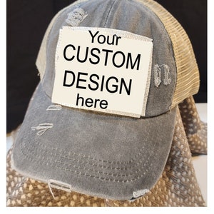 May include: A distressed gray baseball cap with a beige mesh back. The cap features a patch with the text "Your CUSTOM DESIGN here". The hat has a pre-curved bill and a button on top.