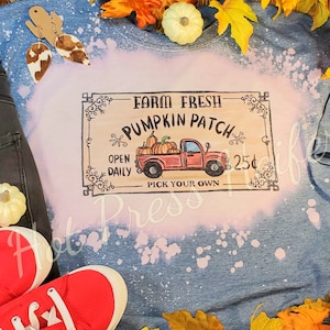 May include: A light blue t-shirt with a bleached design and a graphic that reads "Farm Fresh Pumpkin Patch". The graphic features a red truck carrying pumpkins. Red sneakers and fall foliage are also in the image.