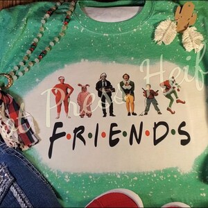 May include: A green and white bleached sweatshirt featuring characters from the movie "Elf" and the word "FRIENDS" in a stylized font. The sweatshirt has a crew neck and long sleeves. A beaded necklace and denim jeans are in the image.