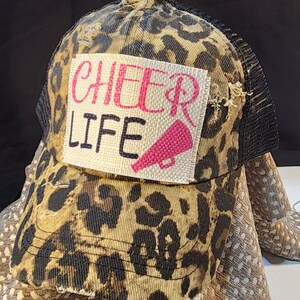 Puede incluir: Gorra de camionero con estampado de leopardo y parte trasera de malla negra. La gorra presenta un parche con las palabras "CHEER LIFE" en rosa y negro, junto con un gráfico de megáfono rosa. La gorra tiene un aspecto desgastado.